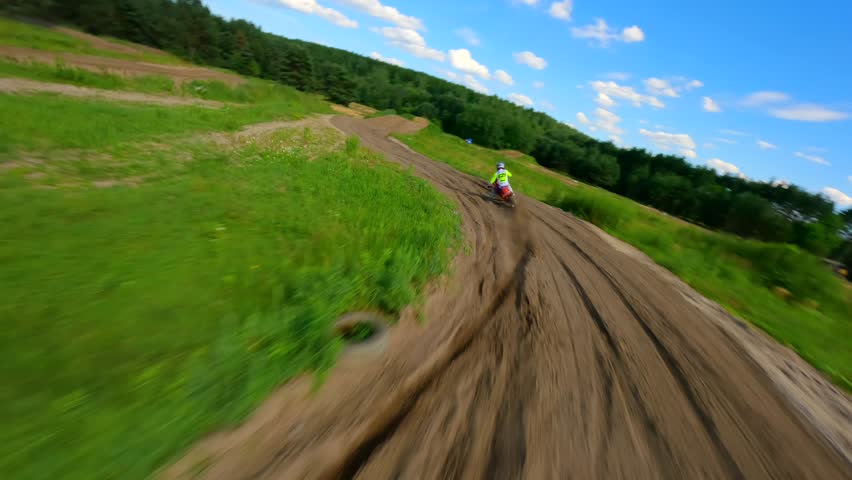 FPV drone chases motocross rider on dirt track through green countryside and forest, capturing fast turns, speed, and outdoor adventure in summer sunlight.
