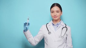 Smiling female doctor in white lab coat and gloves poised with medical syringe, representing vaccination or medical treatment, against light blue background - Powered by Shutterstock - Get 15% off with code: PIKWIZARD15
