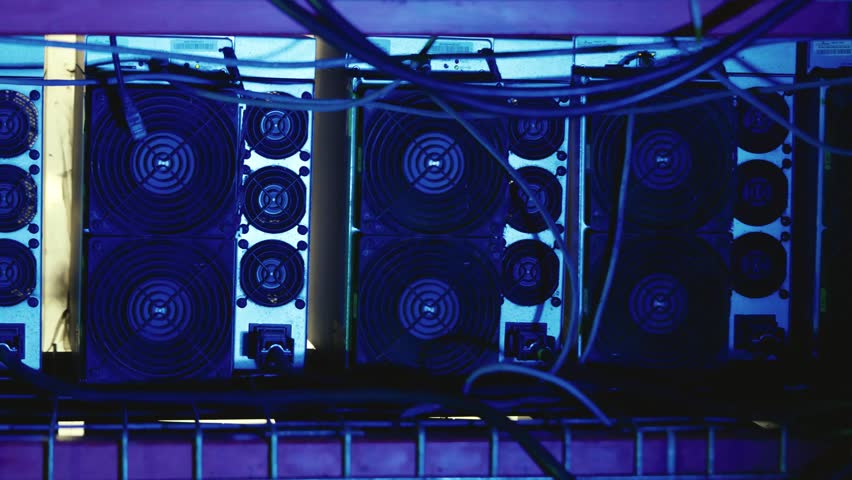 Bitcoin mining rigs with dual cooling fans and connected power and data cables on a metal shelf in a server room. Pan left view