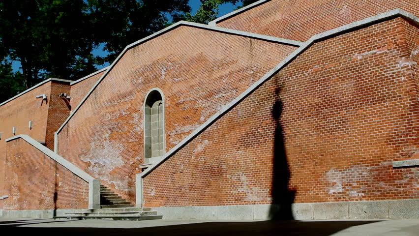 A long red brick wall plays with light and shadow, revealing stairs, textures, and the silhouette of a lamppost in quiet geometry.