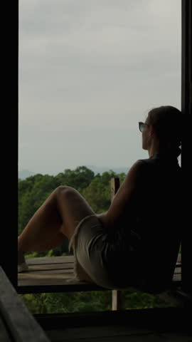 Woman Sitting Alone in Wooden House on Mountain Slope Looking at Sea and Islands Through Open Wall, Backlit Silhouette View, Solitude and Reflection Concept.
