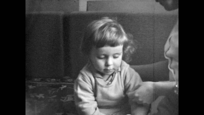 Old Film, Mother undressing child at home. Little girl sitting on couch parent helping to dress. Indoor domestic scene. Daily parenthood life. Vintage family archive. Archival retro 1960s routine
