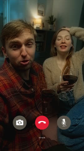 Vertical handheld shot of relaxed man and woman talking on video call over glass of wine in living room when TV suspiciously turning on by itself confusing couple at stormy night