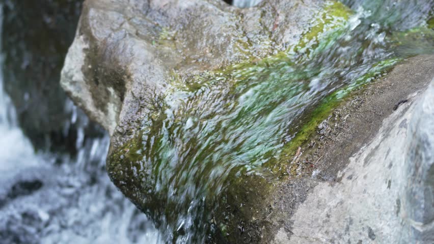 Mossy rock with flowing water. Clear water glides smoothly over a mossy rock surface, creating a soft, natural shimmer. A close-up of nature s quiet movement.