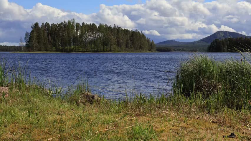 Lake Siljan, Sweden, time-lapse of rippling water and clouds motion in the wind. Green spring grass, Isundaholmen island and in the background Gesundaberget mouintain. Calm northern scenery.