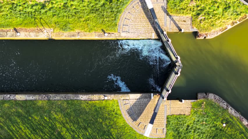 Aerial view canal lock controlling water levels. Overhead shot sluice gate releasing flow. Top-down perspective hydraulic system managing passage. High-angle frame navigational lock adjusting currents