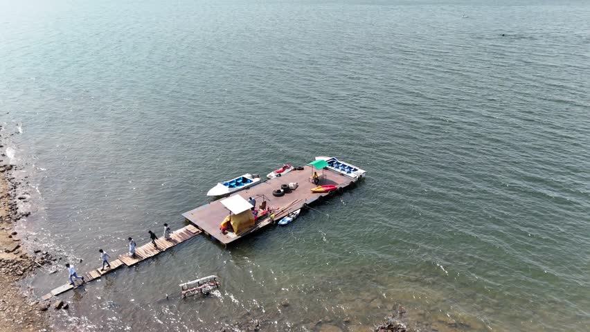 Stunning 4K drone footage showing a large dam with deep blue water in Chhattisgarh, India. Four boats are parked along the side, while six people are seen walking nearby in the scenic environment.

