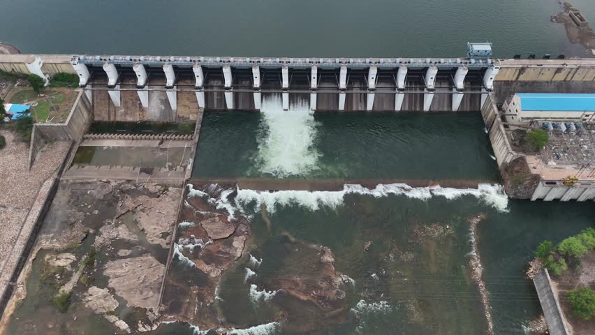 4K aerial drone footage showing the front top view of a fully filled dam in Chhattisgarh, India. Open floodgates are releasing water as the reservoir holds deep blue water under clear skies.