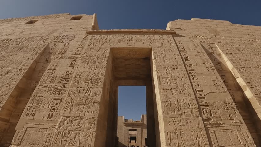 Wide-angle view of the grand stone entrance to an ancient Egyptian temple, adorned with detailed hieroglyphic carvings. The shot captures the symmetry and monumental scale of the historic structure.