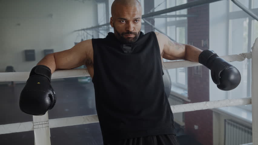 Tired black boxer relaxes leaning on boxing ring ropes after intense training in gym. African American man fighter with gloves expresses strength on arena