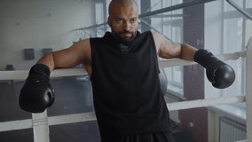 Tired black boxer relaxes leaning on boxing ring ropes after intense training in gym. African American man fighter with gloves expresses strength on arena - Powered by Shutterstock - Get 15% off with code: PIKWIZARD15