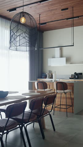 Long wooden table, leather chairs and black mesh pendant under slatted timber roof, flowing to bright minimalist kitchen—perfect establishing shot for upscale villa marketing.