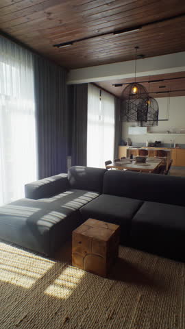 Spacious living room where low afternoon sun streaks across dark sectional, jute rug and kitchen beyond. Earthy palette sells relaxed resort living for promo reels.