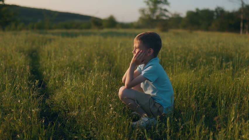 A little boy takes a pause in tall grass with his face in his hands, staring ahead. Surrounded by wild nature, he appears lost in peaceful thought and wonder