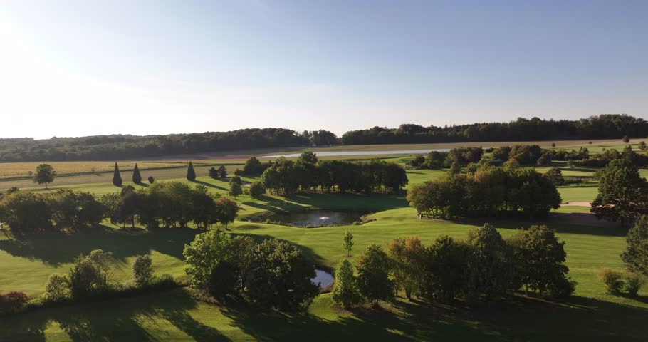 Aerial shot of an idyllic, sunlit golf course with sandbanks and ponds in the middle of the countryside with trees and fields in the background