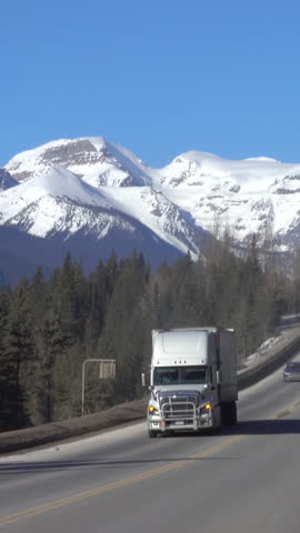 Cinematic shot of freight trucks and cars driving down the road running under the snowy mountains in Banff National Park. Trucks and cars travel across the Canadian wilderness on a sunny winter day.