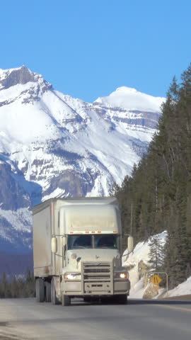 Semi-trailer truck and car drive along the highway crossing Jasper National Park offering a spectacular view of a snow capped mountain range and lush spruce forests. Lorry hauls cargo across Alberta.