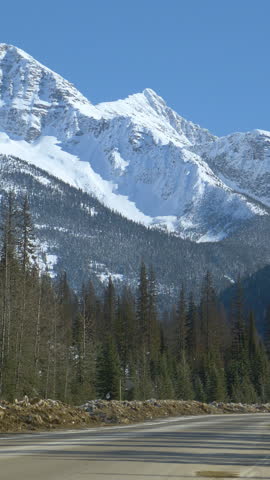 Cars and truck hauling logs drives along road running under snowy mountain range in Alberta. Spectacular snow covered mountains tower above a scenic road leading across the rugged Canadian wilderness.