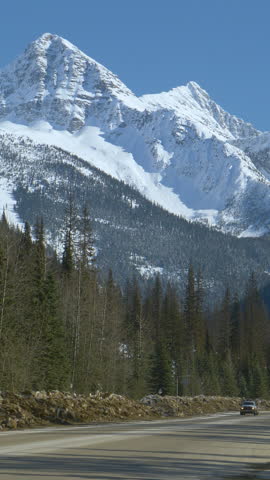 Traffic moves along a scenic country road running under the picturesque snow capped mountains in Alberta, Canada. Cars and cargo truck drive along the Icefields Parkway road running across the woods
