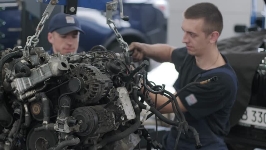 Workers adjusting automotive engine components on hoist.