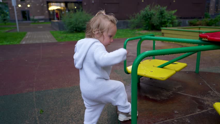 Pensive little girl in white jumpsuit and bandage with broken collarbone and a bandaged arm play on Merry-Go-Round after the rain
