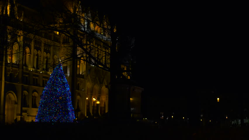 Hungarian parliament building with christmas tree at night. Festive blue-lit christmas tree glowing dramatically near iconic hungarian parliament building, creating luminous winter nightscape
