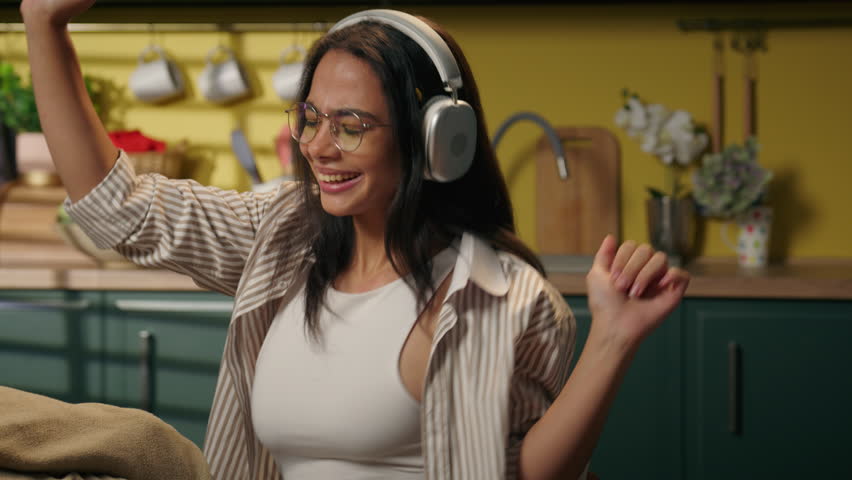 Indian ethnic woman latina girl happy carefree teen female student study freelancer homeowner businesswoman relax at home kitchen listen music headphones enjoy song audio sound in wireless earphones