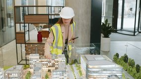 Confident female girl architect in hardhat and reflective jacket using smartphone and analyzing business office complex architectural model. Woman lady smart designer of buildings working in office. - Powered by Shutterstock - Get 15% off with code: PIKWIZARD15