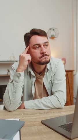 Smart focused man freelancer guy pensive thoughtful sitting at table home office sitting with laptop thinks about solving problem with online service app makes decision deep in thoughts. Vertical