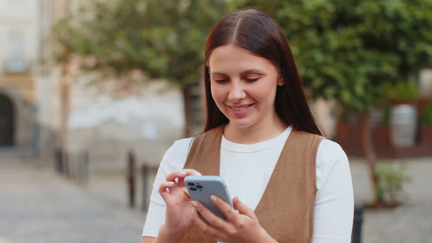 Caucasian young woman using smartphone typing texting social media messages e-mail looking for a way on map in mobile navigator app outdoors. Smiling tourist standing on city street. Town lifestyles.