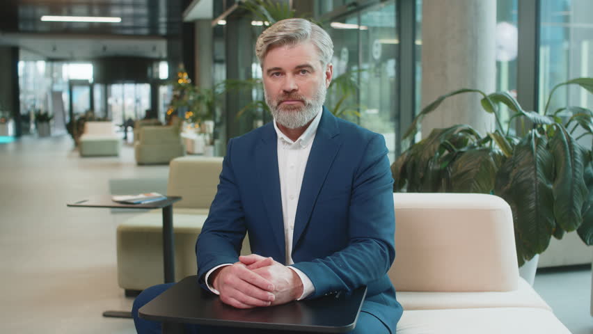 Portrait of mature businessman shakes finger and saying No be careful scolding and giving advice to avoid danger mistake disapproval sign sitting in modern office lobby. Serious guy in suit at hotel.