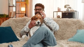 Indian man at home talks on smartphone while beagle dog stays close, listening and resting calmly. Arabian guy on sofa chats during call, showing connection, comfort, warmth, and friendly emotion - Powered by Shutterstock - Get 15% off with code: PIKWIZARD15