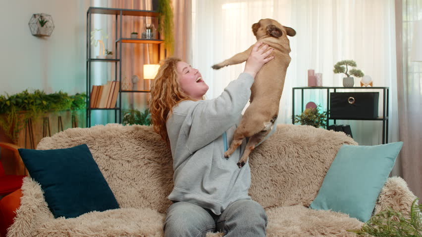 Young Caucasian redhead woman lifts pug dog into air on home sofa, smiling with excitement and victory. Pet waves paws playfully as both express joy in shared energetic celebration moment indoors.