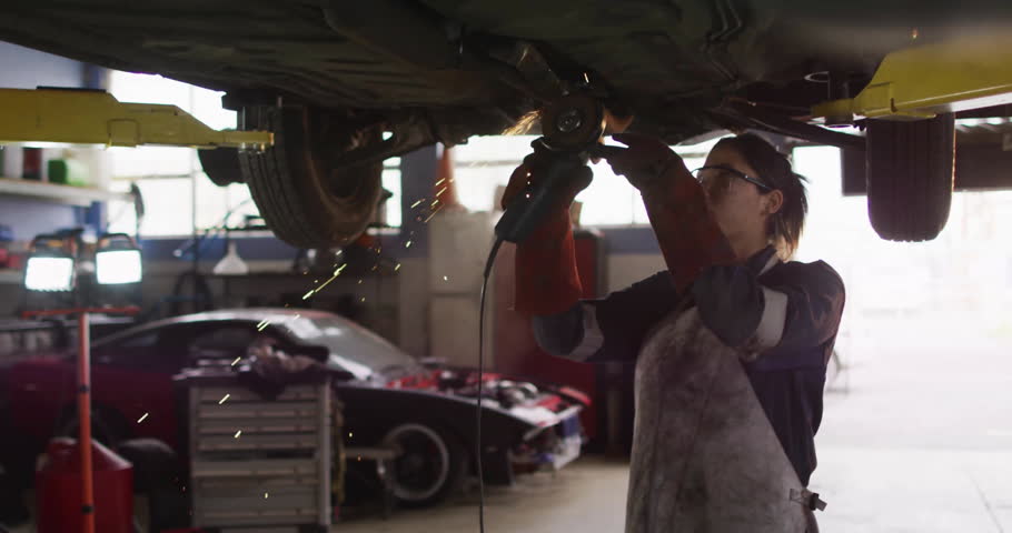 Female mechanic activating angle grinder under auto garage lift, sparking rust removal. Industrial, repair, maintenance, safety, tools, workshop, strength