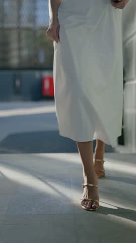 A woman in a white dress and gold high heels walks gracefully on a sunny day. Sunlight and shadows play on her legs as she moves. Her elegant and stylish appearance exudes confidence and femininity.