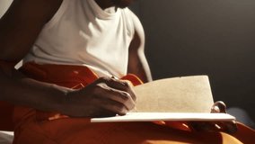 Thoughtful Black inmate in tank top and orange pants writing in notebook on his lap, sitting on bed in prison cell under barred window. Close-up view - Powered by Shutterstock - Get 15% off with code: PIKWIZARD15
