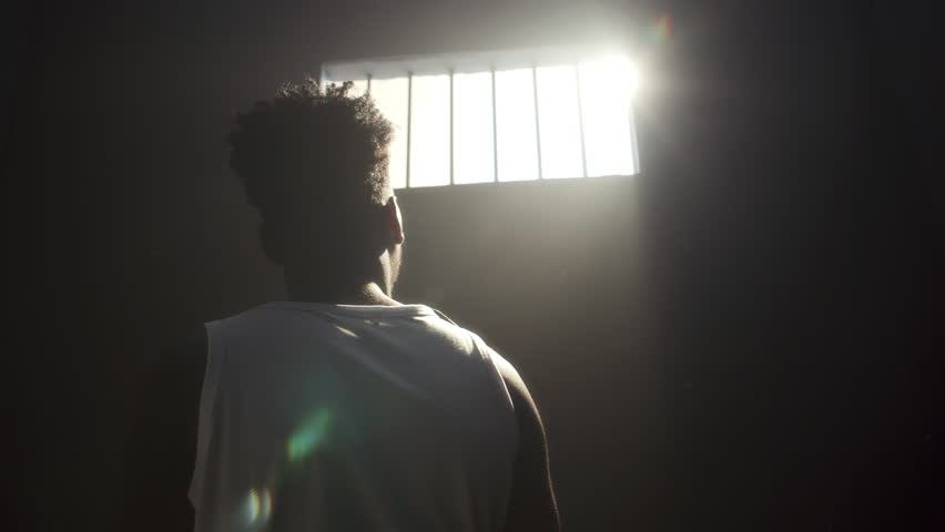 Inmate standing in prison cell with arm extended toward bright sunlight pouring through barred window. View from behind