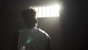 Inmate standing in prison cell with arm extended toward bright sunlight pouring through barred window. View from behind - Powered by Shutterstock - Get 15% off with code: PIKWIZARD15