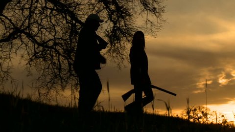 A pair of hunters, in silhouette, walk with their guns, at sunset.