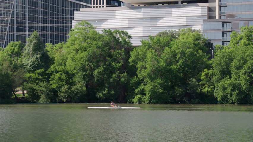 Austin, Texas rower glides across Lady Bird Lake past downtown buildings and thick greenery. The moment captures urban fitness and still water beneath a sunny summer sky in the city center.