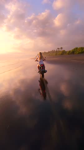 FPV drone shot of woman riding motorcycle along tropical beach during golden sunset, creating cinematic, free-spirited atmosphere. Concepts of freedom, adventure, and slow travel lifestyle.