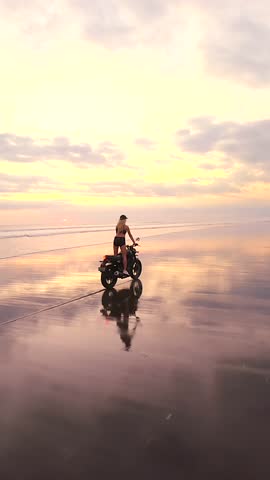 FPV drone shot of woman riding motorcycle on wide black sand beach in Bali at sunset in slow motion. Cinematic scene symbolizing freedom, exploration, and slow travel lifestyle.