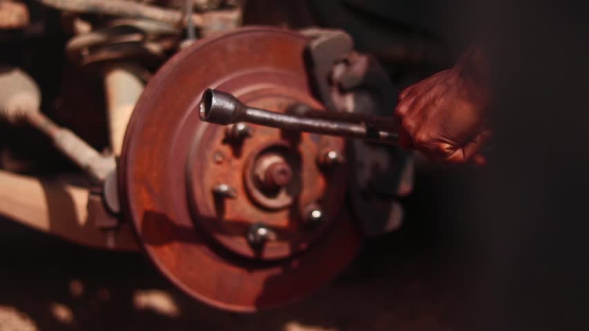 Fixed camera close-up of a mechanic’s hand using a tool on a rusted car brake in Nigeria.