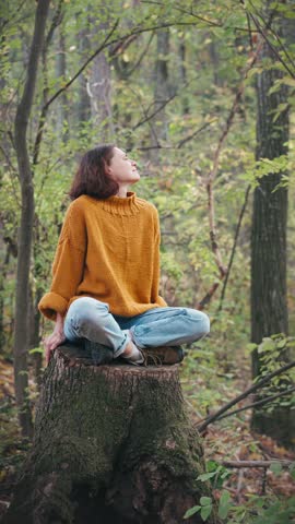 A young woman enjoys the fresh air while sitting on the stump of a large tree in the autumn forest