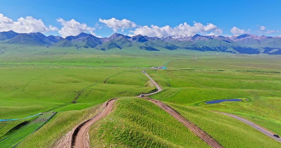 Aerial shot of a winding road through green grasslands and majestic mountain range under blue sky in Xinjiang, China.