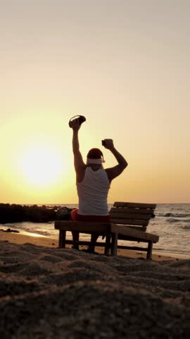Santa Claus dances. Funny Santa Claus dancing with a wireless musical bluetooth speaker in hand, against backdrop of sunset or sunrise. back view. relax on beach by the sea