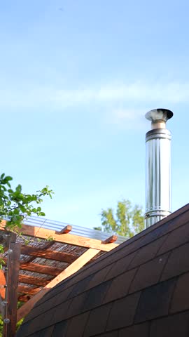 Colorful golden hour vertical outdoor closeup of traditional Nordic Scandinavian Estonian sauna house hut rooftop with narrow clean metal chimney and white smoke calmly exiting as sauna is prepared 