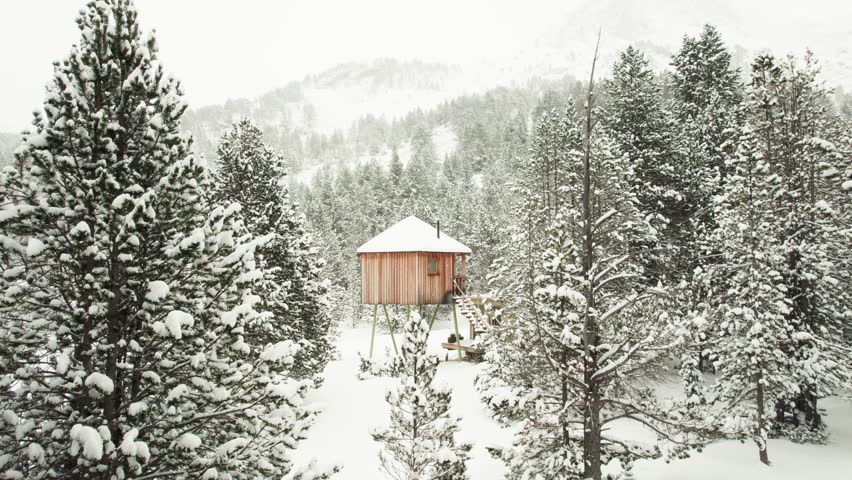 Drone flies through snowy trees toward stilted wooden cabin in Andorra, with snowfall and mountain backdrop.