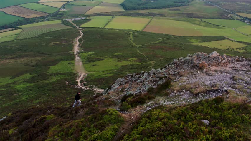 Standstill drone shot of a man climbing the Sugar Loaf’s rocky trail with green fields below during golden evening
