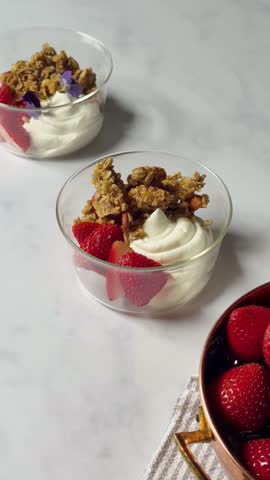 A vertical footage of a hand adding edible flowers in yogurt with granola and strawberries on a marble countertop